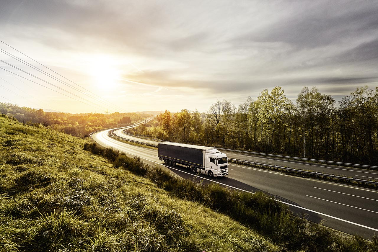 904377080 White truck driving on the highway turning towards the horizon in an autumn landscape with sun shining through the clouds in the sky